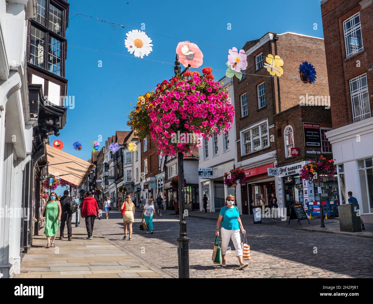 Guildford High Street con alcuni acquirenti che indossano maschere facciali. Cesti floreali autunnali e colorati striscioni stagionali in chiaro blu caldo sole Foto Stock