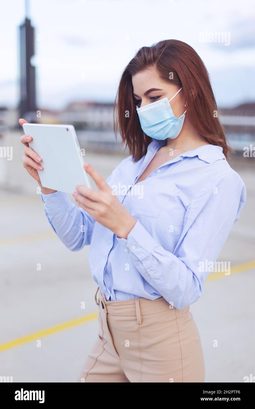 Giovane donna d'affari caucasica 20s in maschera utilizzando tablet all'aperto in città Foto Stock