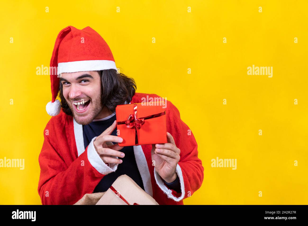 L'uomo sorridente vestito come Babbo Natale mostra un piccolo regalo rosso preso da un grande sacco pieno di regali. Sfondo giallo e spazio di copia per il vostro g Foto Stock