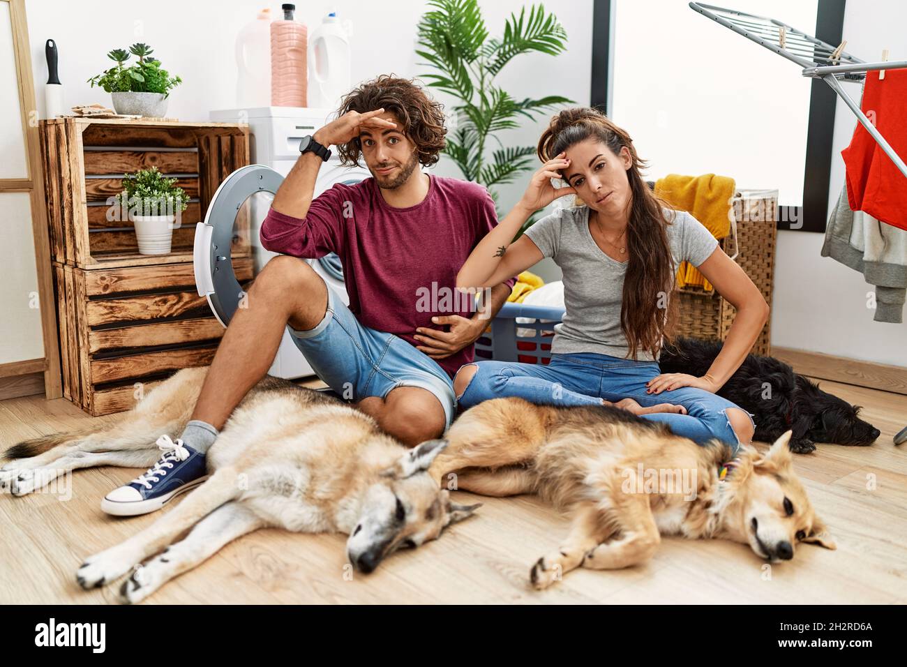 Giovane coppia ispanica che fa la lavanderia con i cani preoccupati e sollecitati circa un problema con la mano sulla fronte, nervoso e ansioso per la crisi Foto Stock