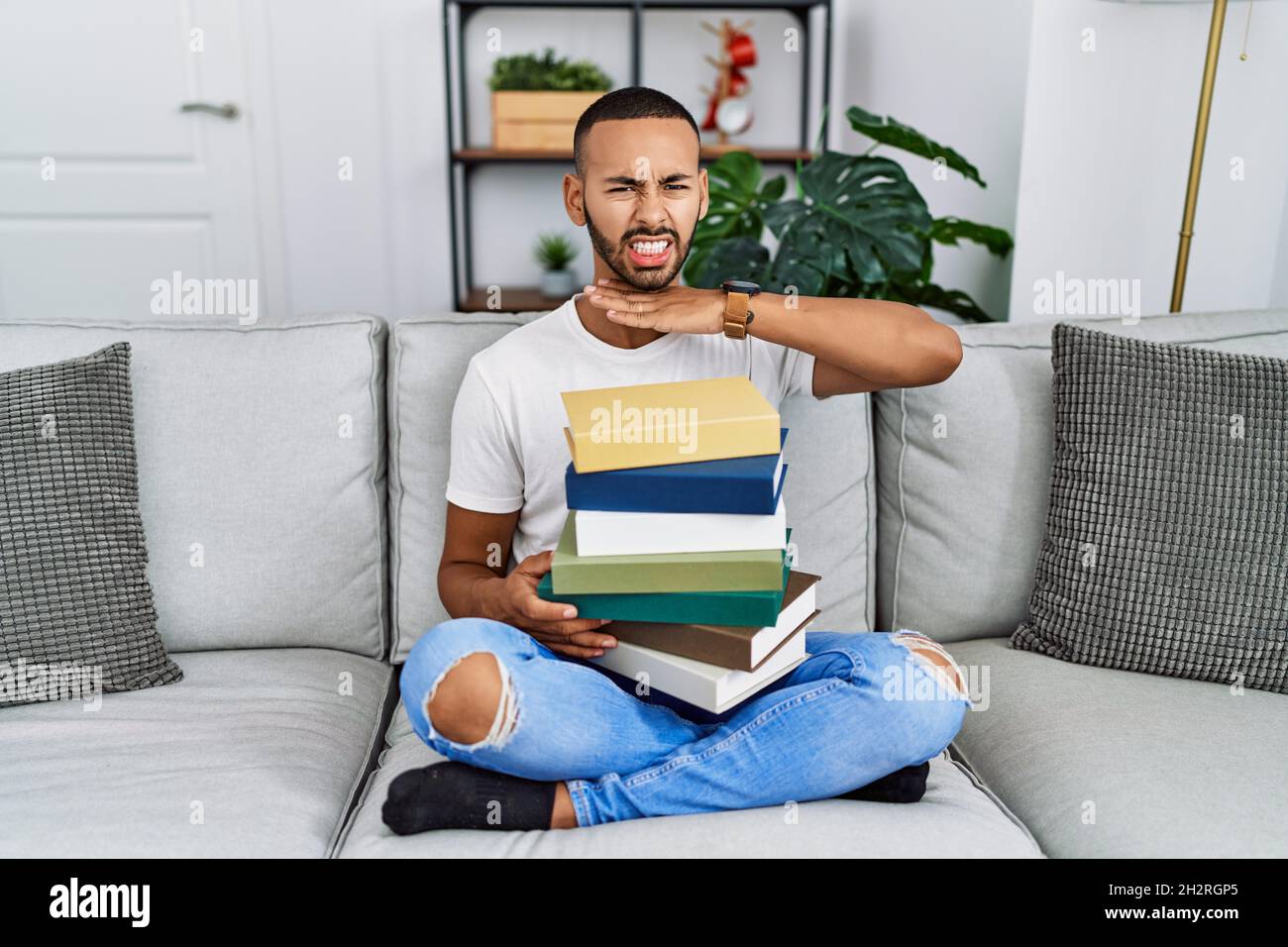 Il giovane afroamericano che tiene un mucchio di libri seduti sul divano tagliando la gola con la mano come coltello, minaccia l'aggressione con violenza furiosa Foto Stock