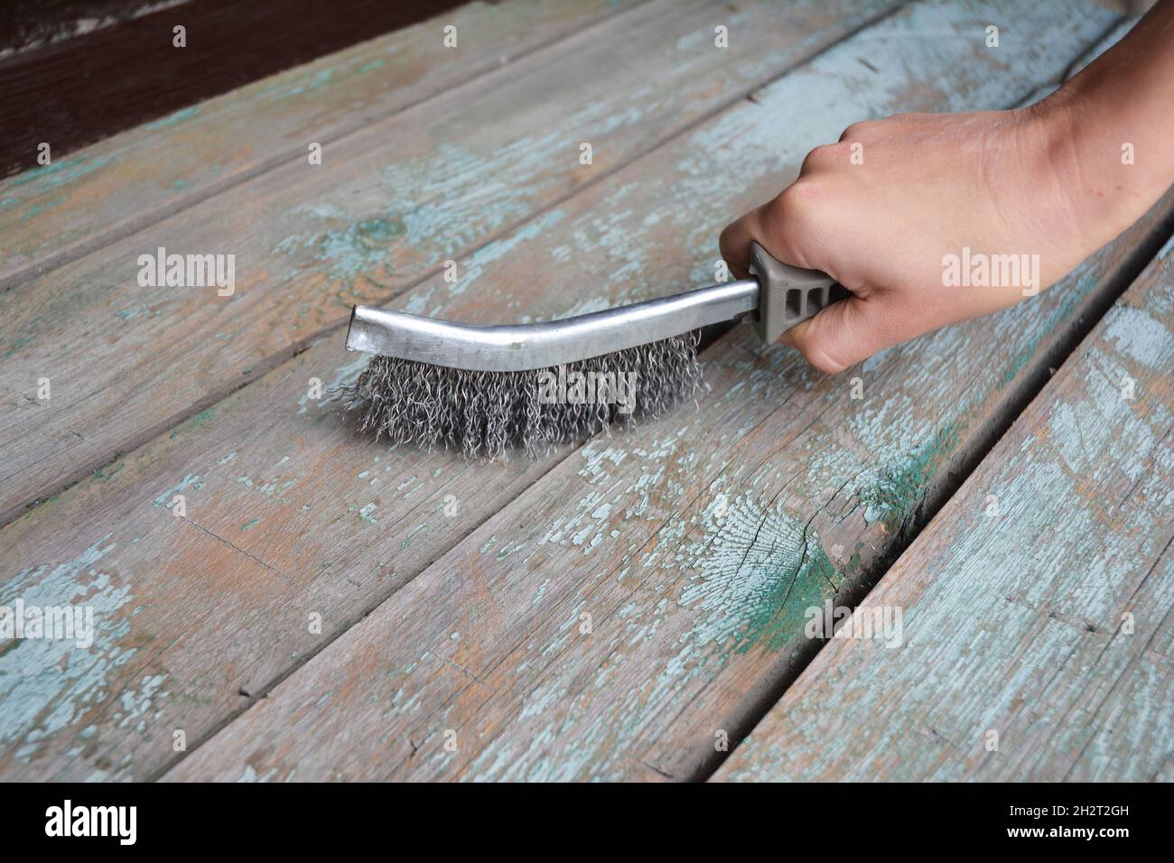 Un uomo sta rimuovendo la vecchia vernice dalla superficie di legno con una spazzola metallica. Foto Stock