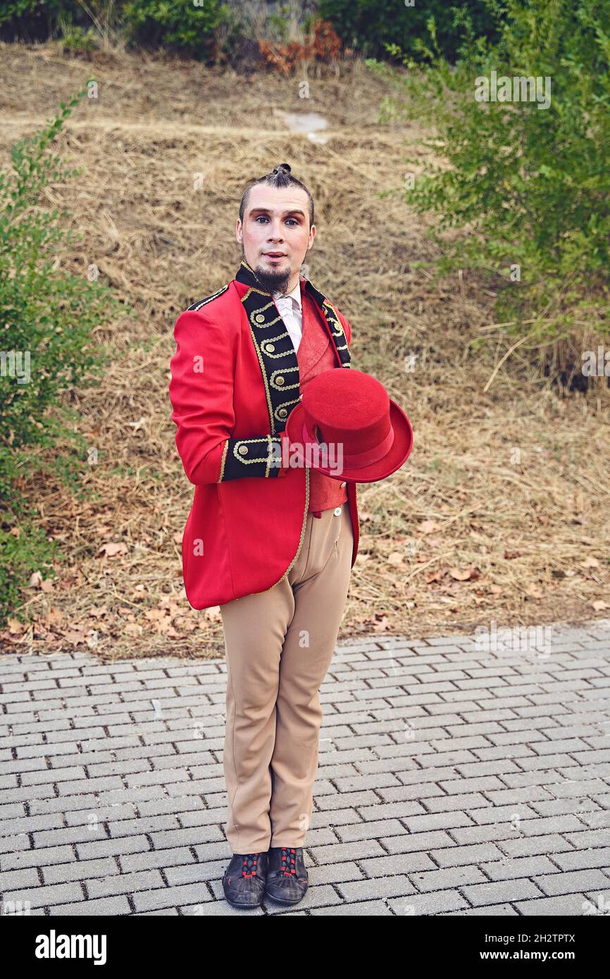 Uomo a corpo pieno in costume elegante che tiene il cappello e guarda la macchina fotografica mentre si trova in piedi sul marciapiede durante lo spettacolo nel parco Foto Stock