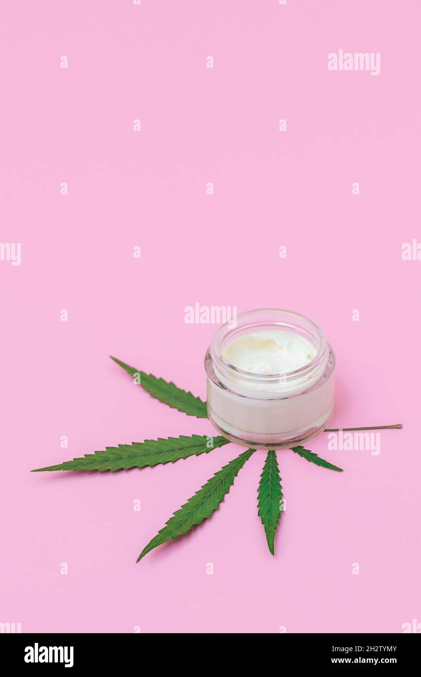 Vaso in vetro cosmetico con crema facciale infusa di cannabis e foglia verde di marijuana su sfondo rosa pastello, immagine verticale Foto Stock