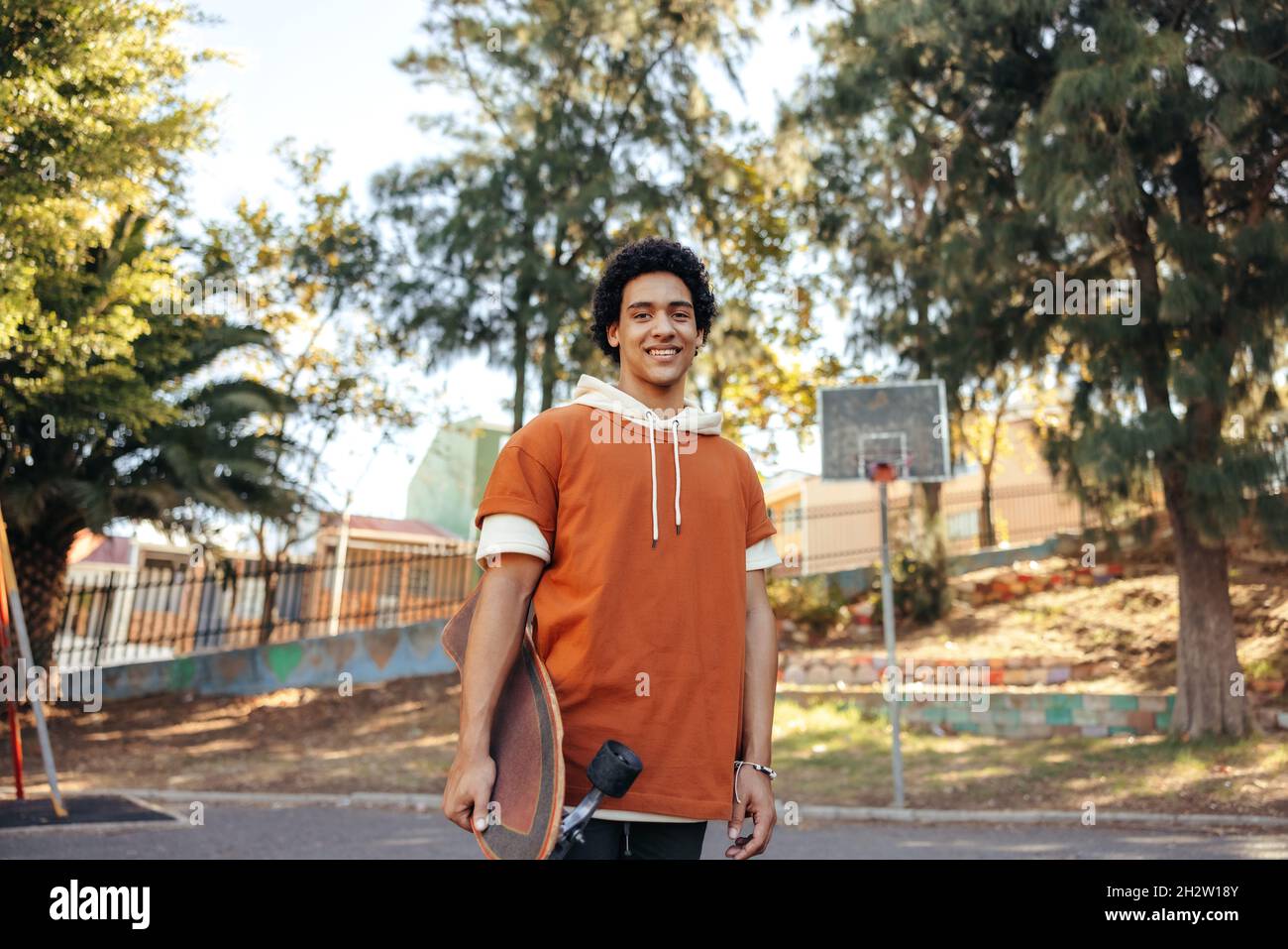 Skateboarder felice che trasporta il suo skateboard all'aperto durante il giorno. Allegro adolescente sorridente alla telecamera mentre è in piedi all'aperto. adolescente sportivo Foto Stock