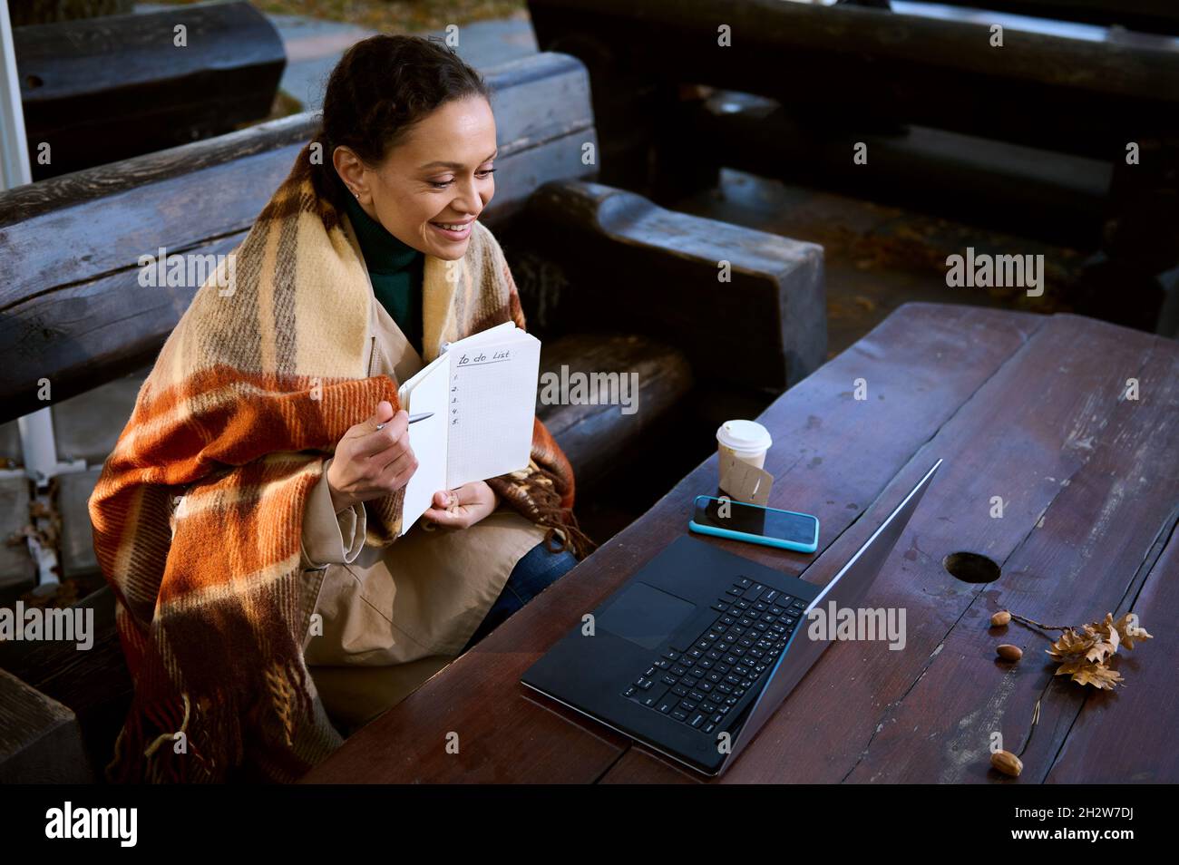Sorrisi donna attraente mostrando con penna su una pagina vuota con elenco di cose da fare durante la videoconferenza su un laptop, si mantiene caldo avvolgendosi in lana Foto Stock