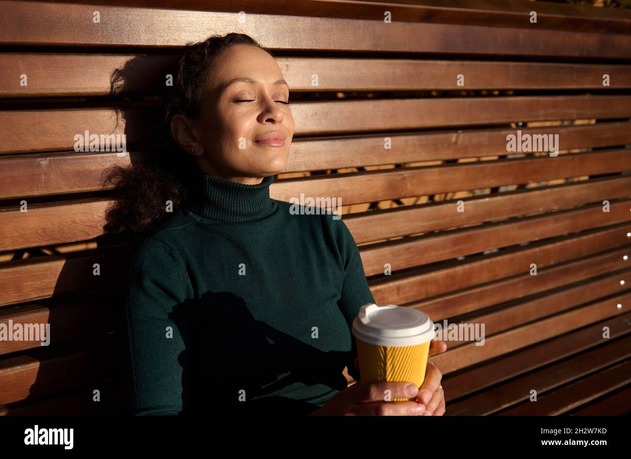 Primo piano di una giovane donna tranquilla e affascinante seduta su una panca di legno, appoggiata sulla schiena in un parco, tiene una tazzina di carta da asporto con bevanda calda restin Foto Stock