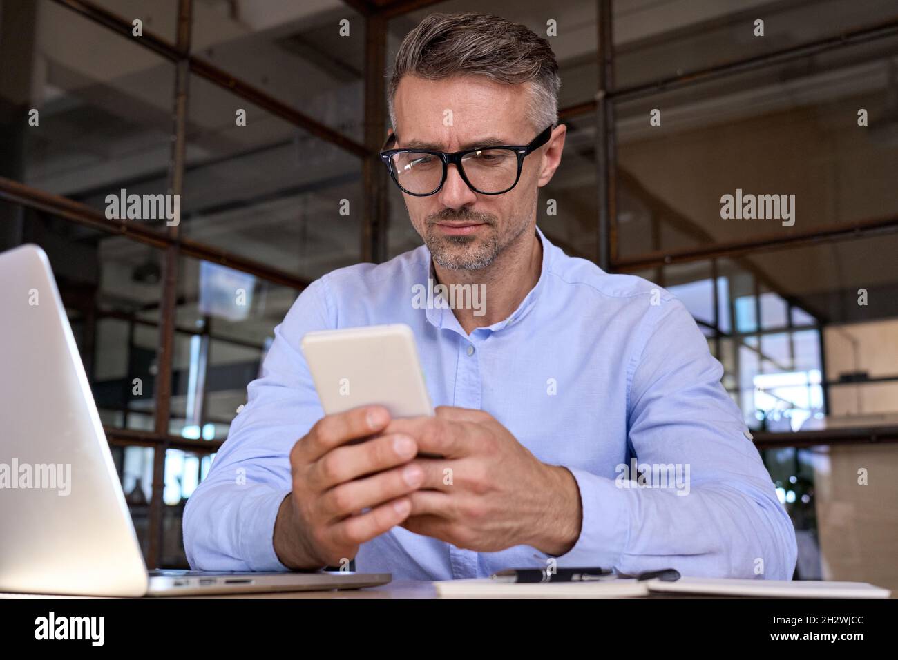 Uomo d'affari maturo che tiene la cellula usando il telefono che controlla i dati che funzionano in ufficio. Foto Stock