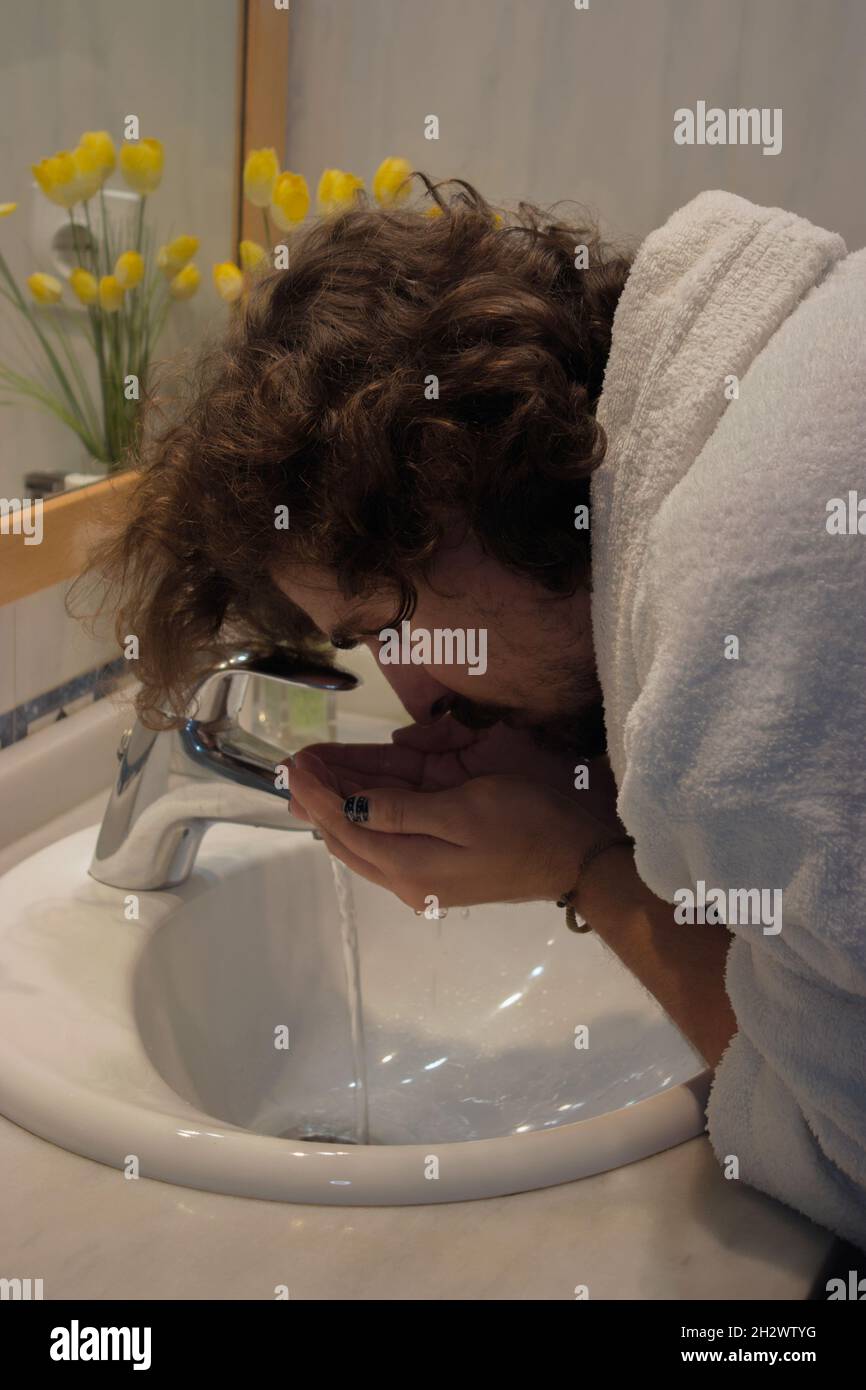 Il giovane in accappatoio si lava il viso nel lavandino del bagno prima di radersi. Home Spa Concept Beauty Concept. Foto Stock