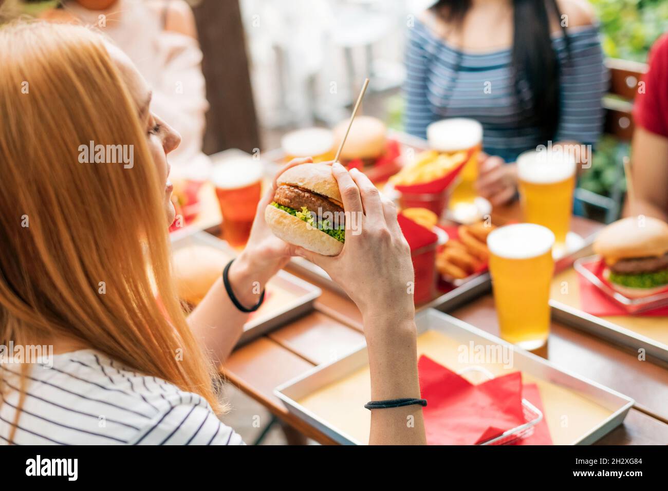 Vista laterale della giovane donna che si prepara a bocciare l'hamburger gustoso intero durante il pranzo con gli amici al tavolo con spuntini e bevande nel ristorante Foto Stock