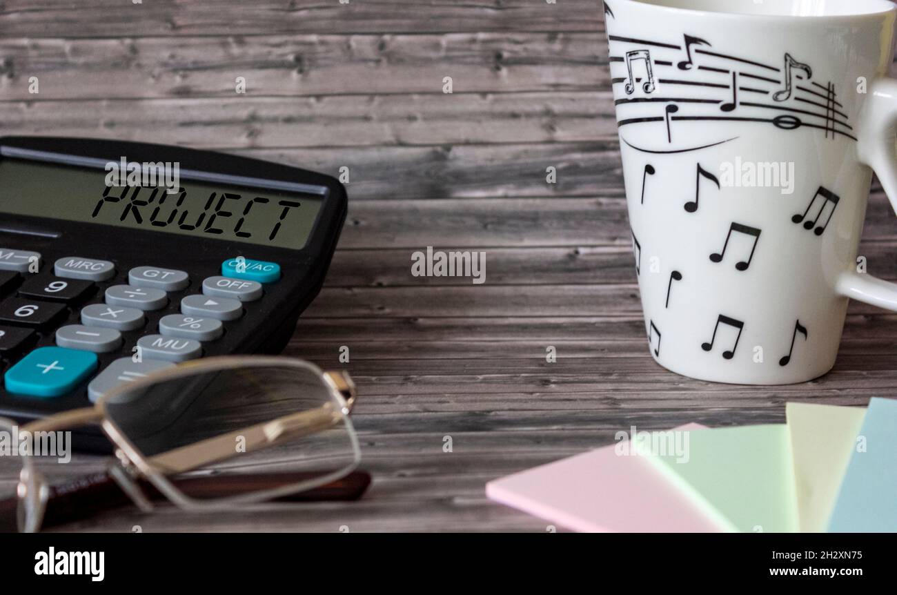 Su un tavolo di legno si trova una calcolatrice con il testo progetto, adesivi, occhiali e una tazza Foto Stock