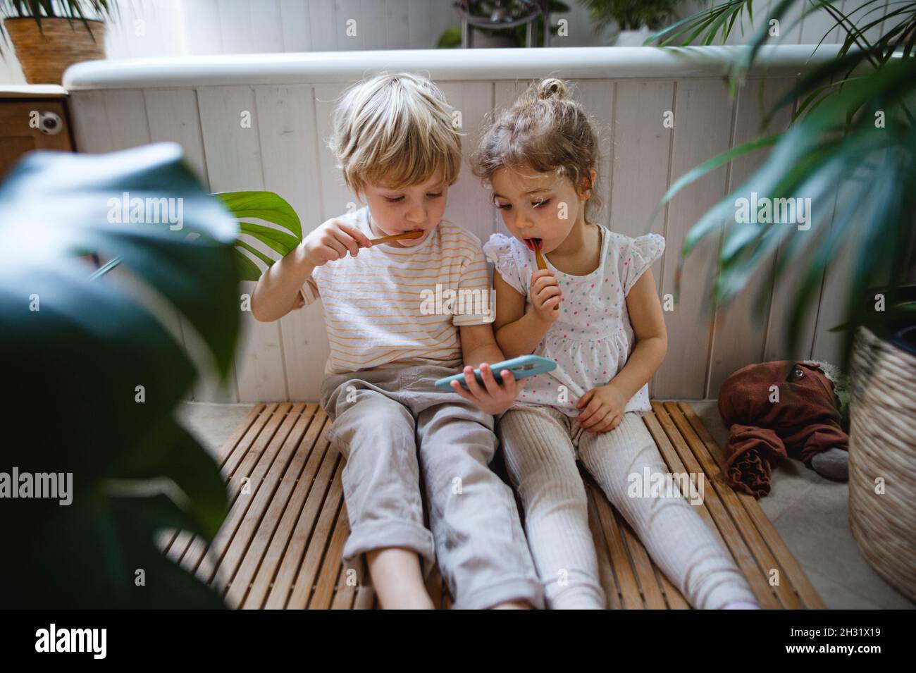 Bambini piccoli e carini seduti sul pavimento in bagno, spazzolando i denti e utilizzando lo smartphone. Foto Stock