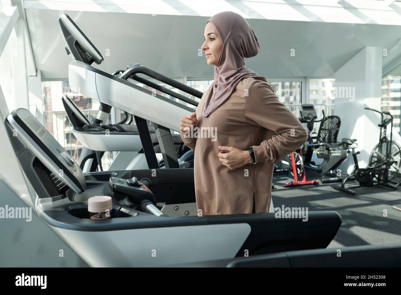 Giovane donna in hijab e abbigliamento sportivo che si esercita su una grande varietà di macchine sportive in grande palestra moderna Foto Stock