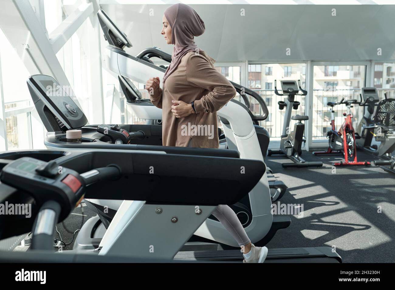Giovane donna piuttosto musulmana in abbigliamento sportivo e hijab in esecuzione sul tapis roulant in palestra contro varie attrezzature sportive Foto Stock