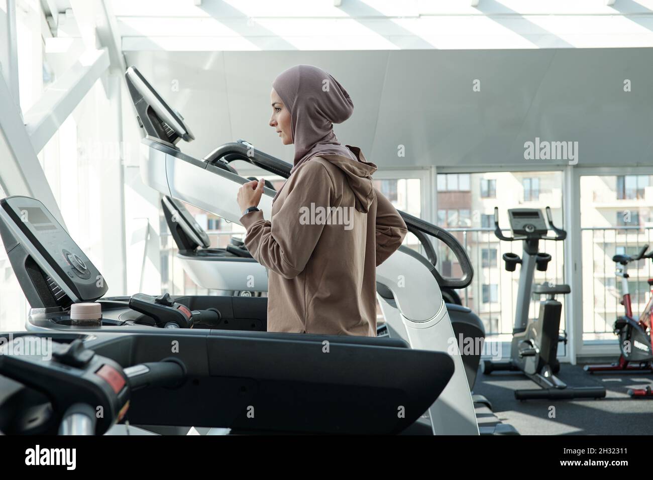 Giovane donna attiva in hijab e abbigliamento sportivo che corre sul tapis roulant mentre passa il tempo libero in palestra Foto Stock
