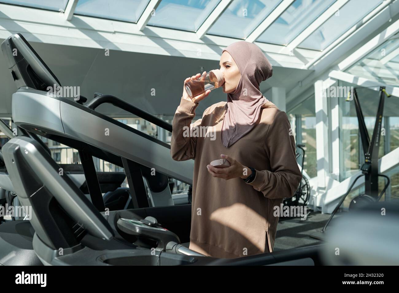 Giovane donna musulmana che ha acqua dopo l'addestramento duro sul tapis roulant nella grande palestra contemporanea Foto Stock