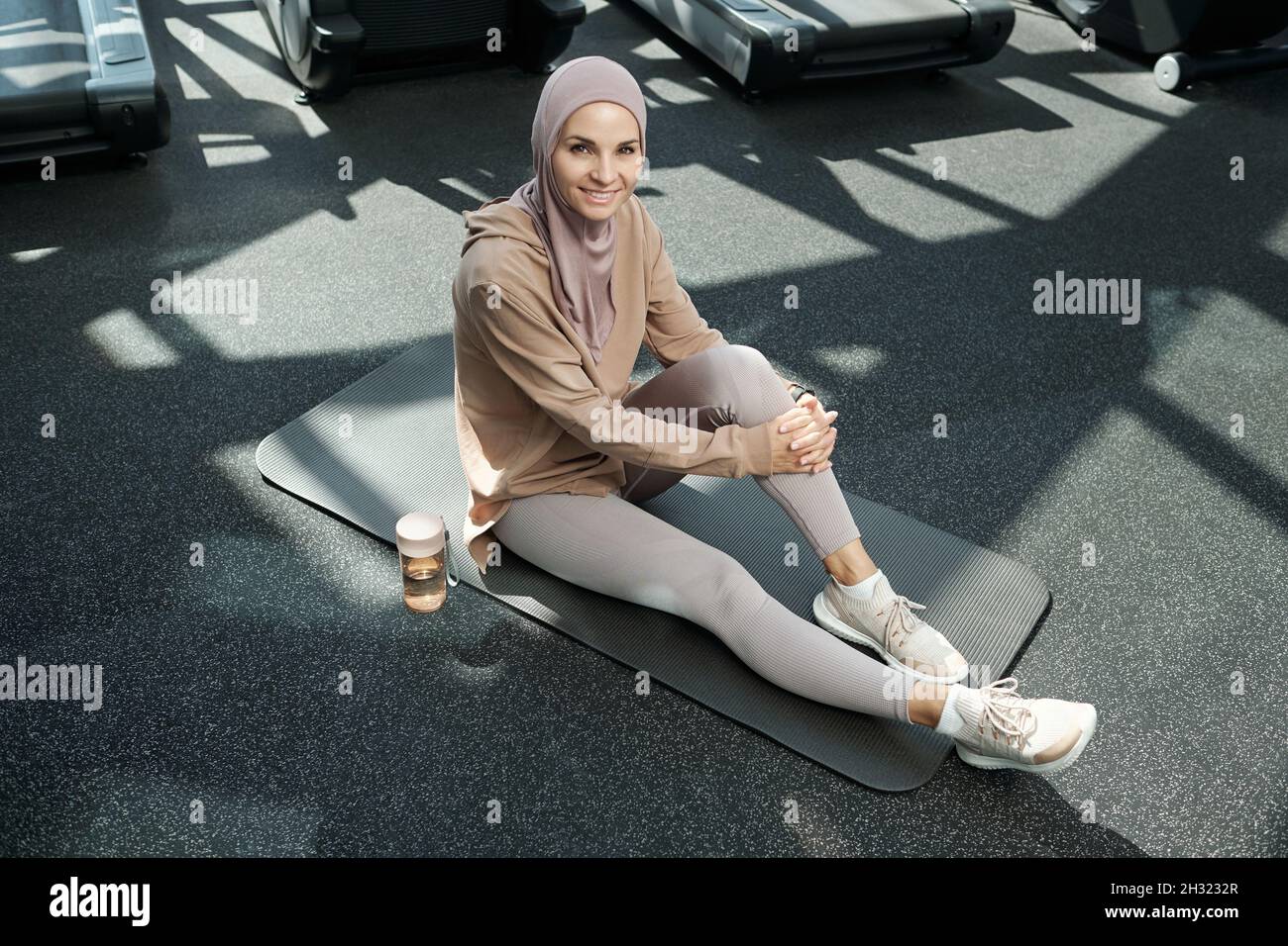 Felice giovane sportivo in hijab e abbigliamento sportivo che ti guarda mentre sei seduto sul tappeto in palestra Foto Stock
