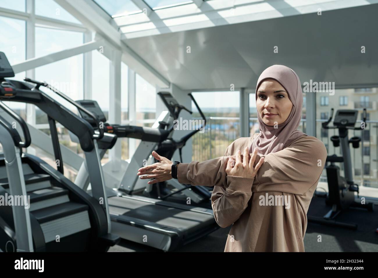 Giovane sportivo in hijab facendo esercizio fisico per le braccia mentre si alza in palestra tra le attrezzature sportive Foto Stock