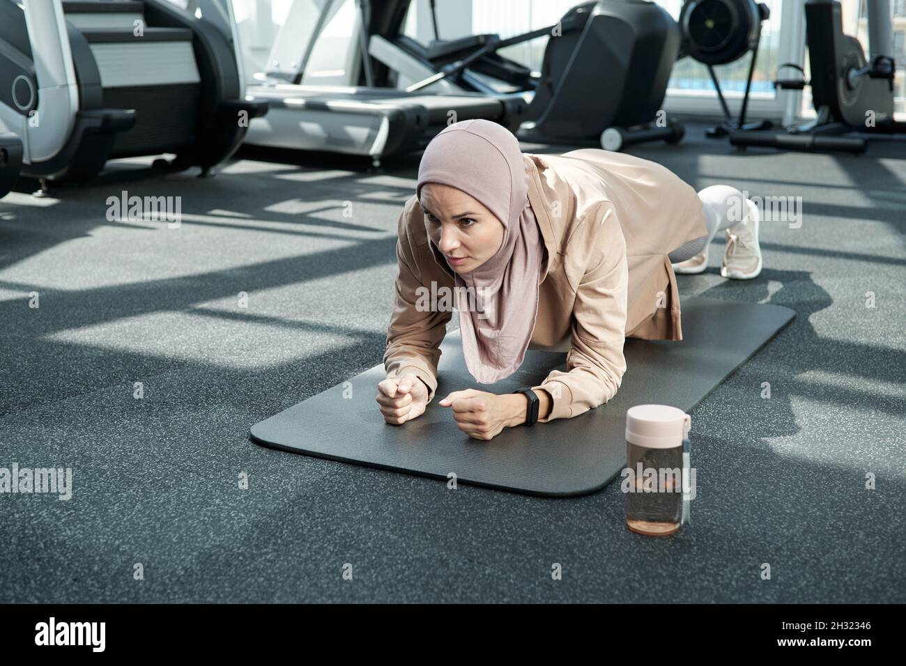 Giovane donna in hijab e abbigliamento sportivo facendo fatica mentre si fa l'asse sul tappeto nel centro fitness o in palestra Foto Stock