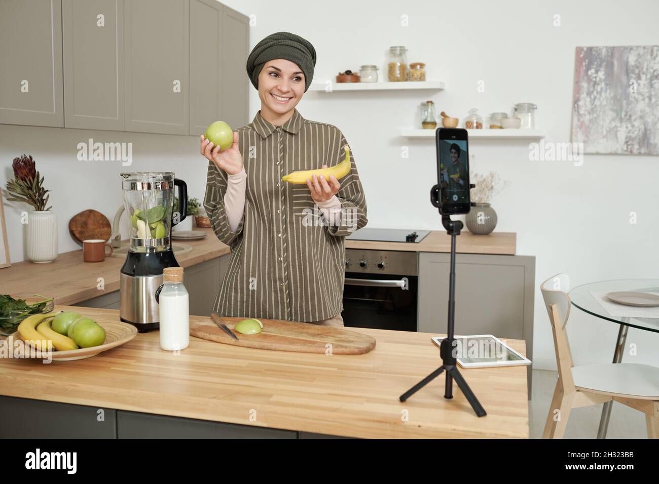 Giovane donna sorridente che mostra banana fresca e mela al suo pubblico online mentre prepara frullato di frutta fatta in casa Foto Stock