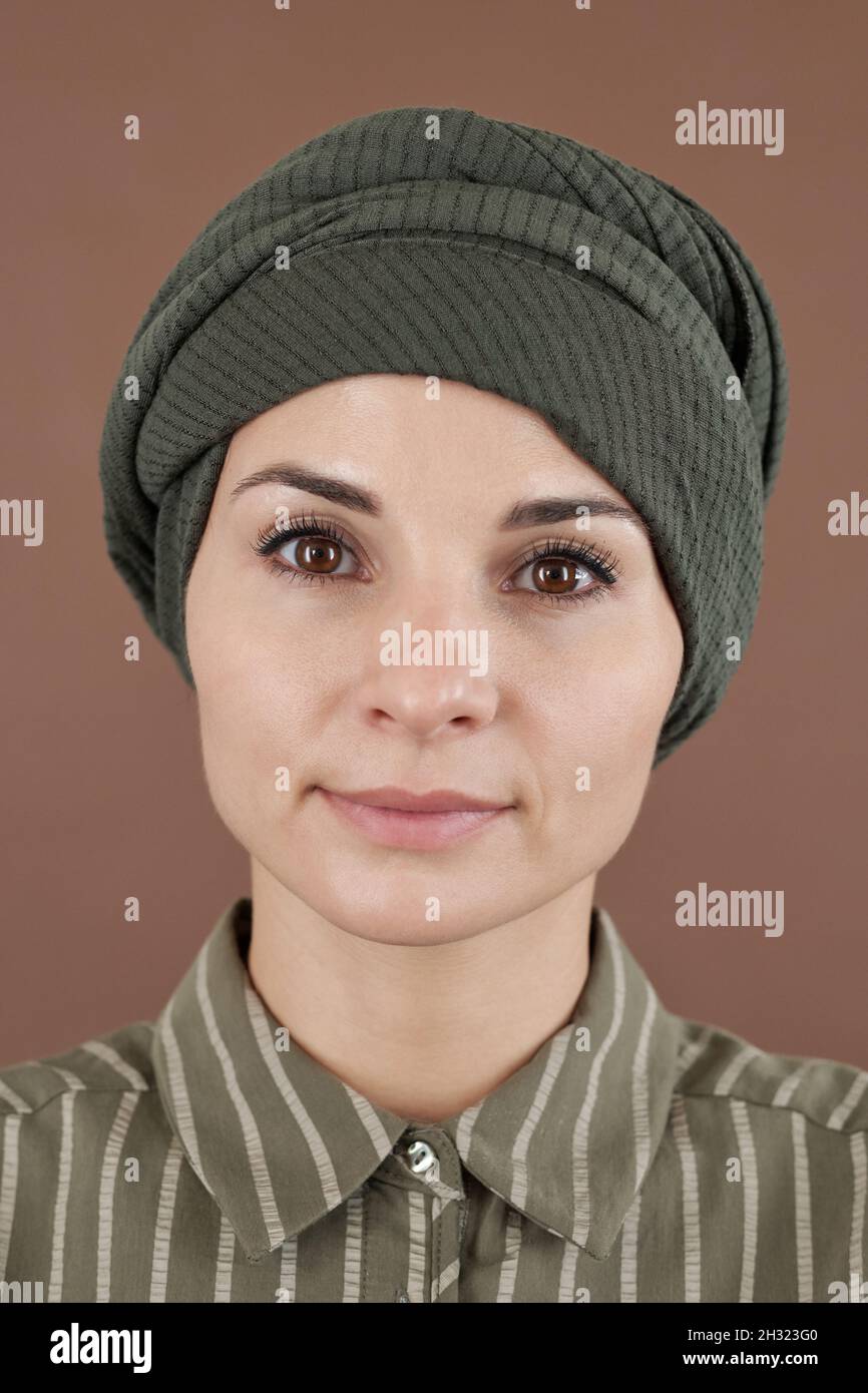 Faccia della giovane femmina musulmana in headwear e camicia a righe in piedi davanti alla macchina fotografica e guardando voi Foto Stock