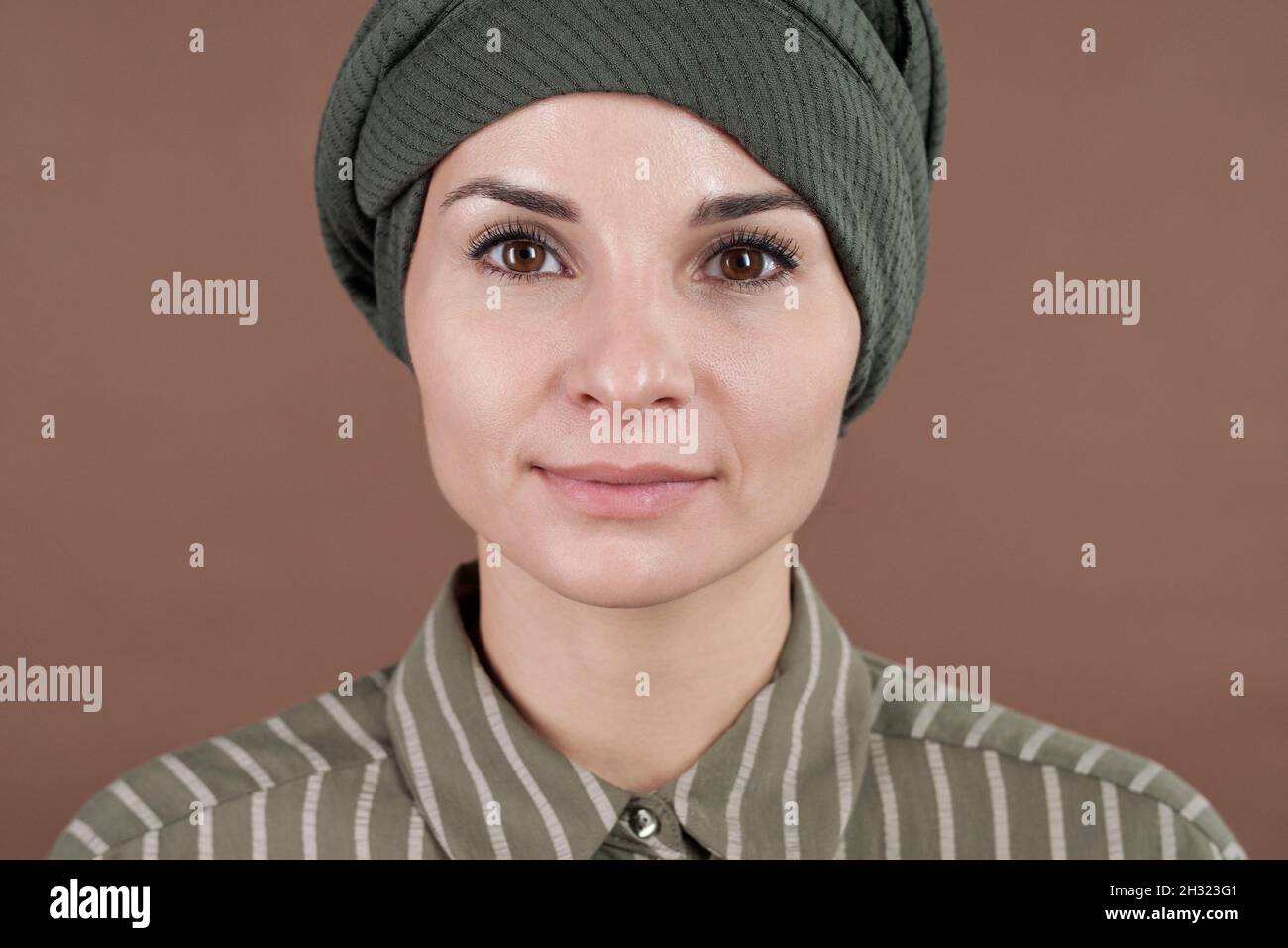 Faccia di giovane donna musulmana contemporanea seria in maglia a righe e headwear guardando la macchina fotografica Foto Stock