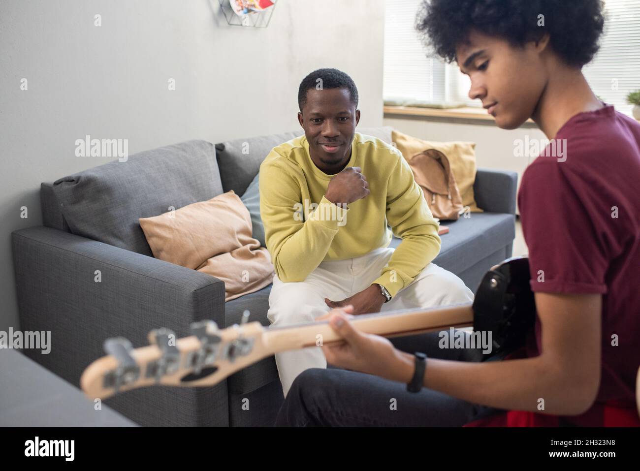 Happy blackman in casualwear guardando il ragazzo giovane che suona la chitarra durante la lezione di musica in ambiente domestico Foto Stock