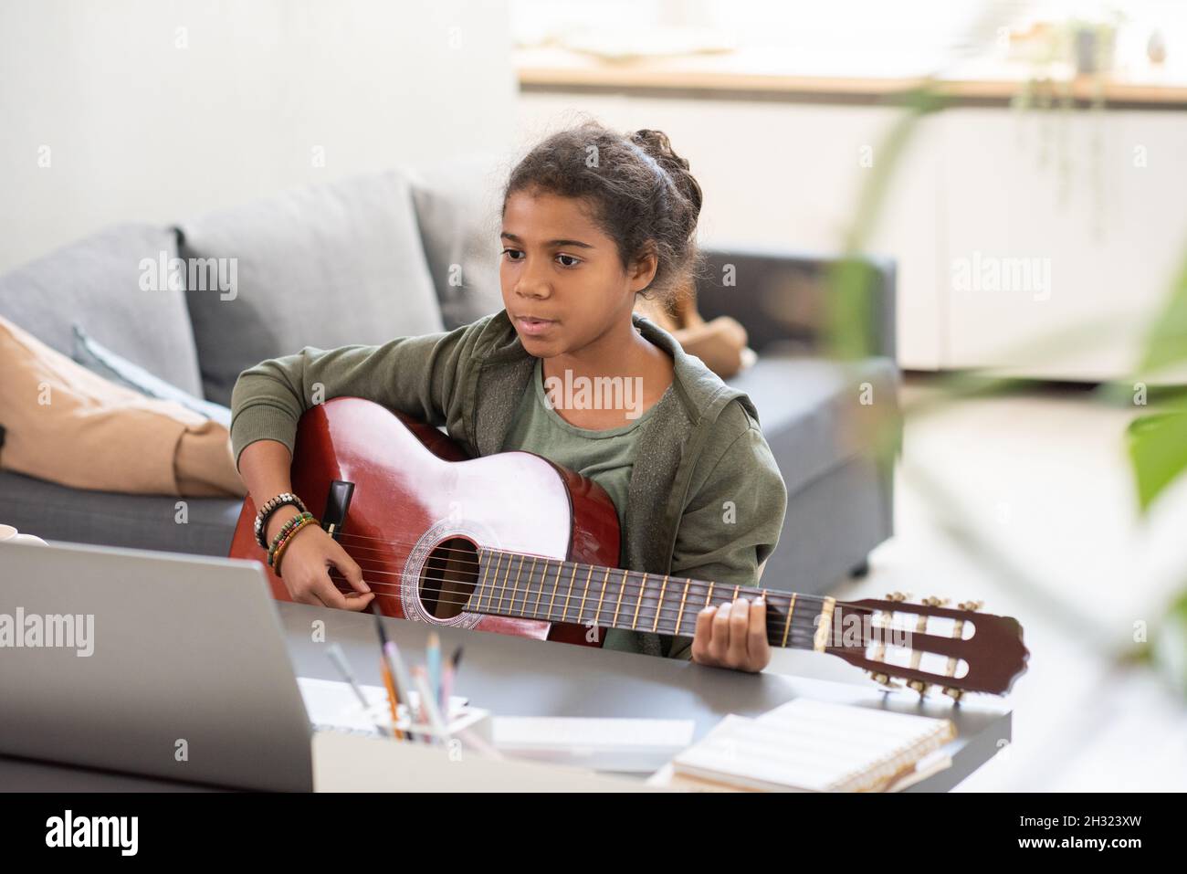 Studentessa seria con chitarra seduta da tavolo di fronte al portatile e ascoltando il consiglio dell'insegnante durante la lezione online Foto Stock