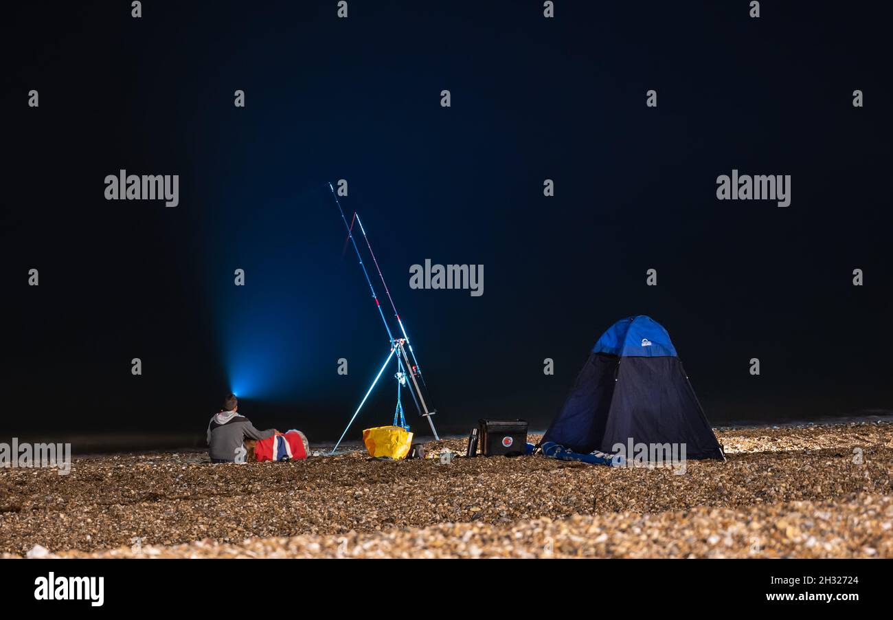 Una giovane coppia con faro su una spiaggia di ghiaia di notte, pesca in mare o oceano con una canna da pesca e tenda. Preso dopo il buio in Inghilterra, Regno Unito. Foto Stock