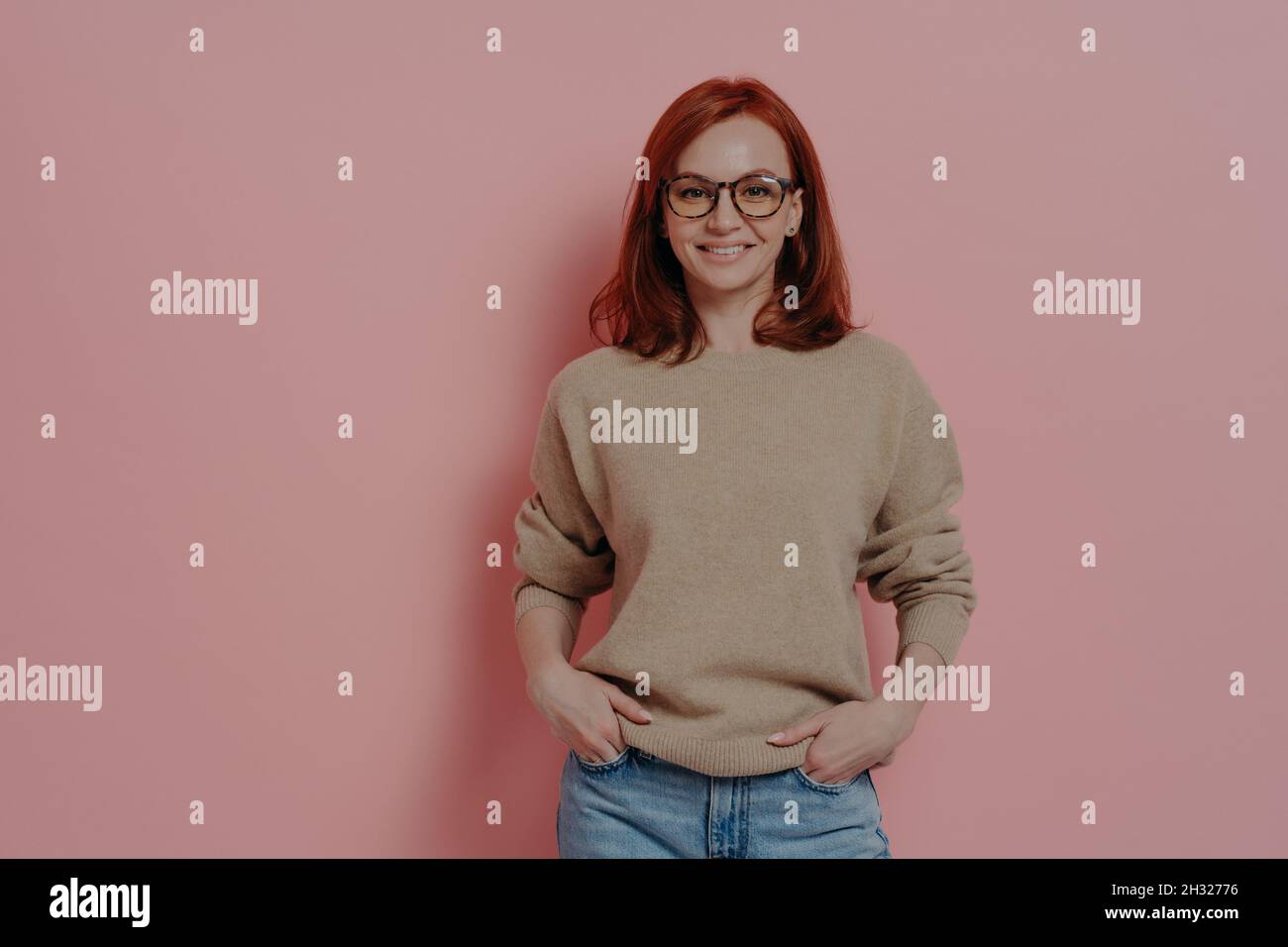 Felice studentessa rossa in occhiali sorridenti alla macchina fotografica, isolato su sfondo rosa Foto Stock