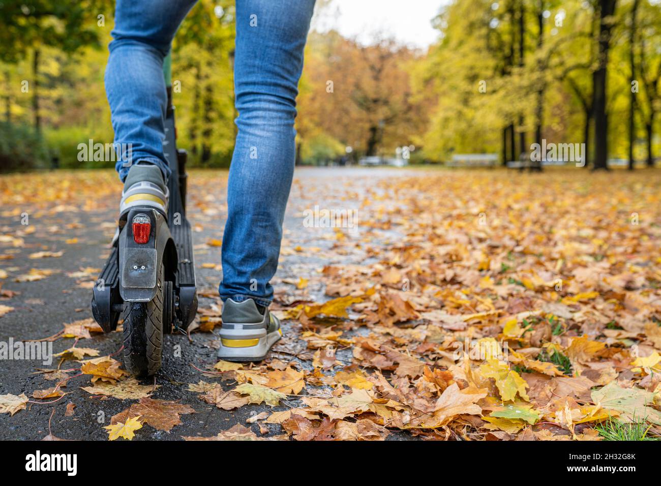 Gambe di un uomo in jeans e sneakers in piedi su scooter elettrico sul marciapiede in autunno strada con spazio copia. Persona irriconoscibile su uno scooter e-scooter cittadino moderno. Concetto di noleggio scooter elettrico Foto Stock