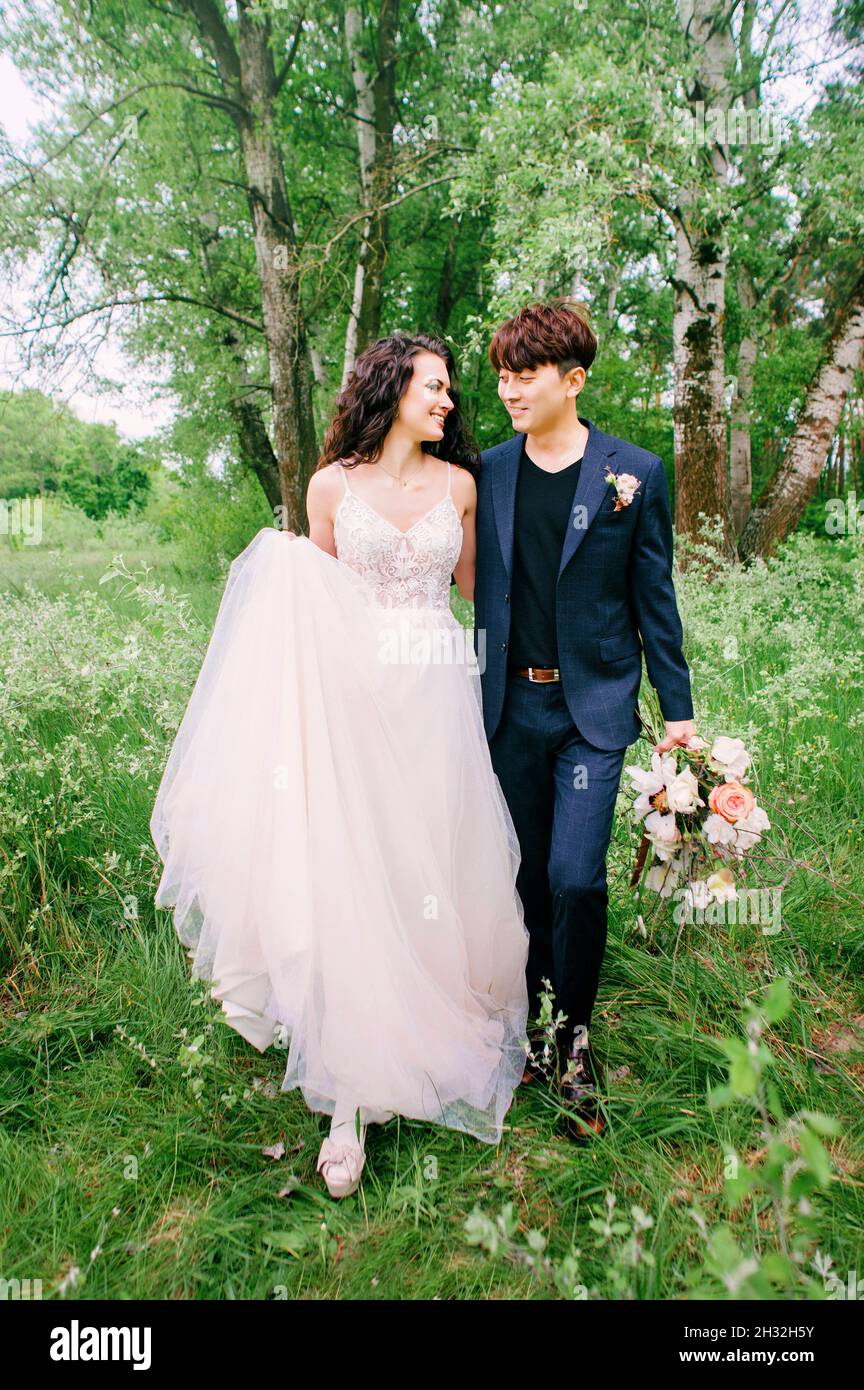 Sorridenti sposi che camminano e si divertono nella fresca foresta verde. Ritratto di sposa e sposo. Cerimonia di nozze all'aperto di primavera. Internazionale sposato co Foto Stock