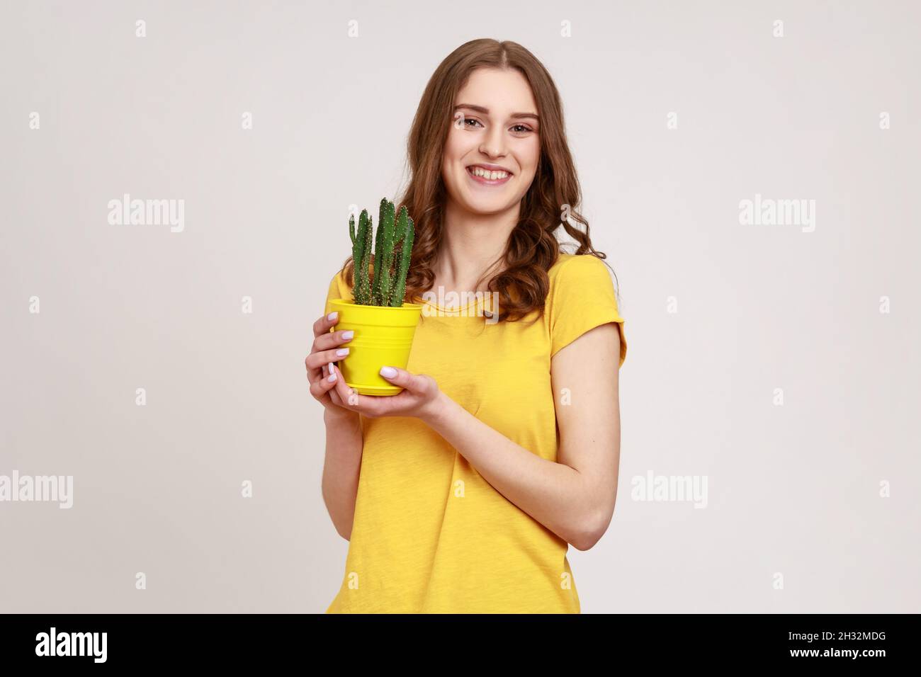 Cura per la pianta in vaso. Ritratto di giovane donna in T-shirt gialla con vaso di fiori e cactus, guardando la macchina fotografica con un sorriso toothy. Studio interno girato isolato su sfondo grigio. Foto Stock