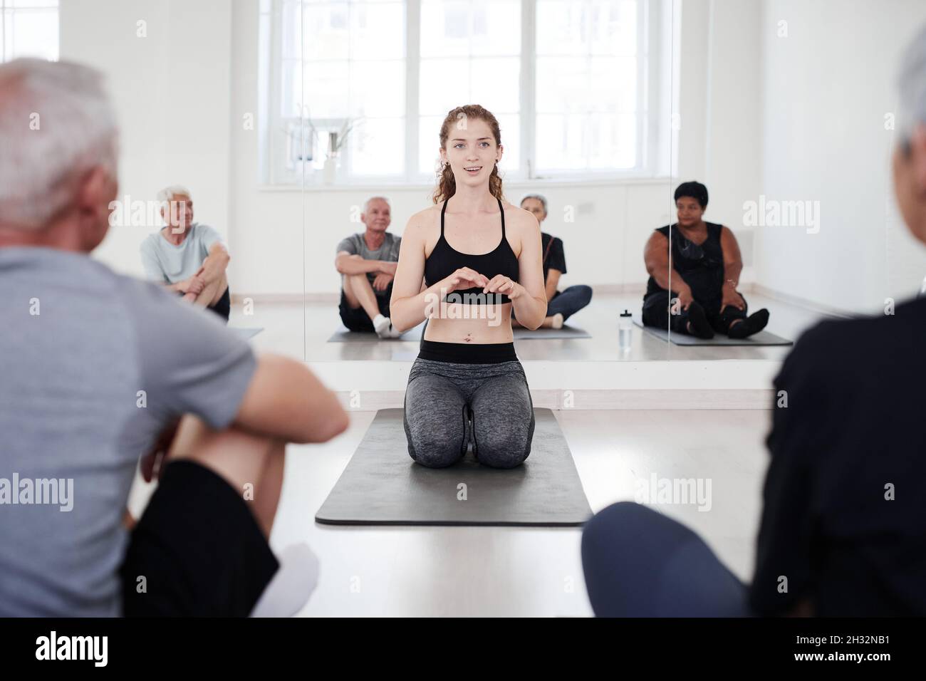 Giovane donna sana seduta sul materassino da esercizio e parlare con gli anziani durante la lezione di yoga in studio Foto Stock