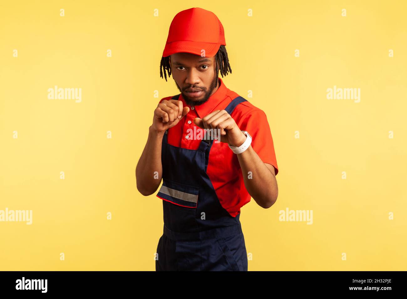 Ritratto di uomo da lavoro aggressivo con barba che indossa uniforme mantenendo i pugni aggrappati, pronto per la lotta, professione di manutenzione della pulizia. Studio interno girato isolato su sfondo giallo. Foto Stock