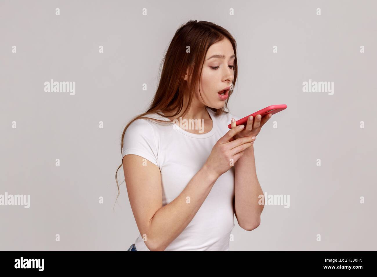 Donna attraente che tiene il telefono cellulare con entrambe le mani e parla in altoparlante di carico, facendo nota vocale, assistente vocale, indossando una T-shirt bianca. Studio interno girato isolato su sfondo grigio. Foto Stock