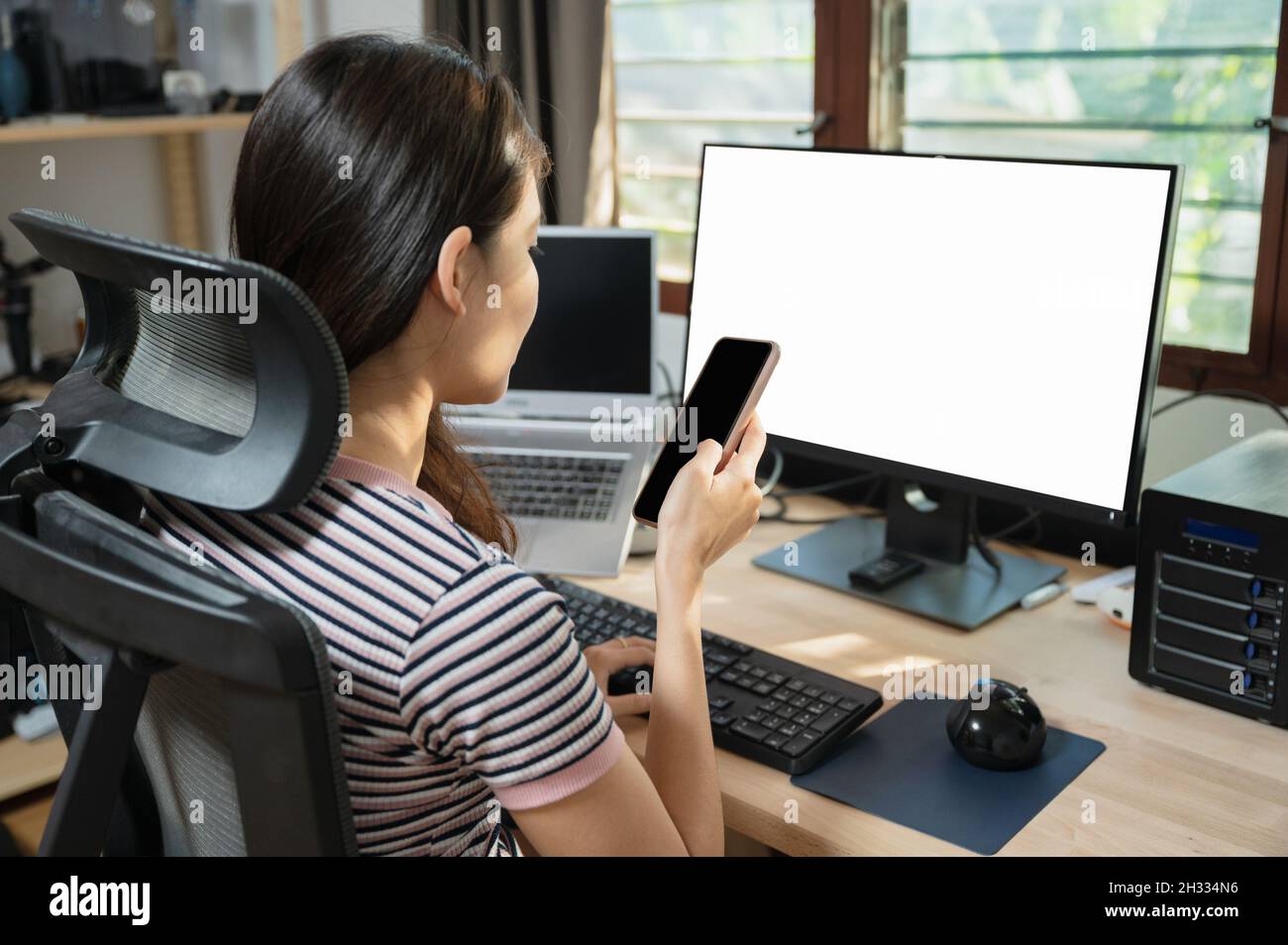 Ragazza asiatica freelance che usa il telefono cellulare al tavolo di lavoro con monitor vuoto in casa Foto Stock