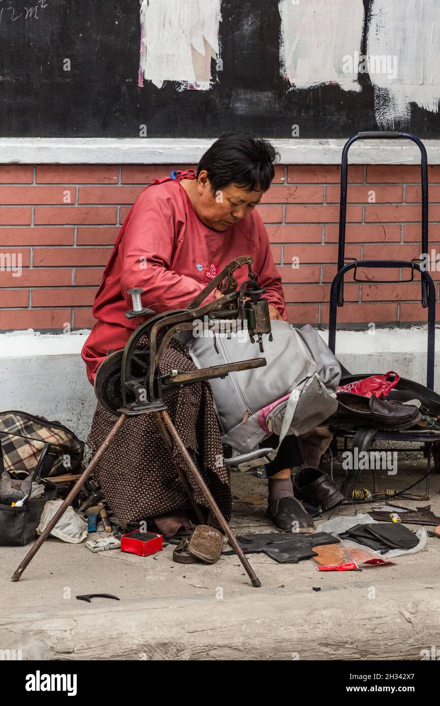 Una donna ripara uno zaino con la sua macchina da cucire manuale sulla strada nella Città Vecchia, Shanghai, Cina. Foto Stock