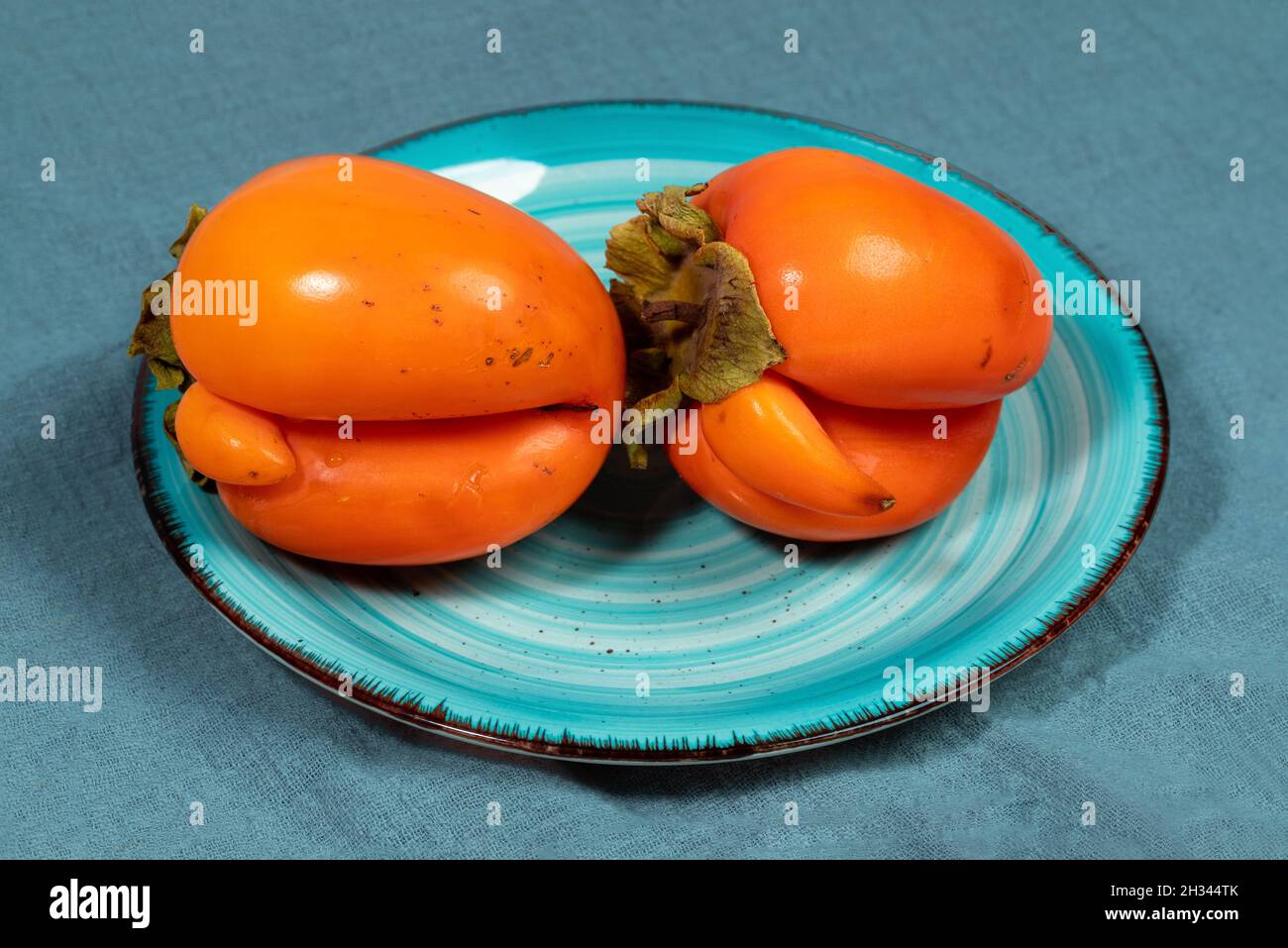 Piatto turchese con due brutti persimmoni arancioni su tovagliolo in tessuto turchese scuro da vicino. Frutta fresca sana biologica. Concetto di cibo senza rifiuti. Reas Foto Stock