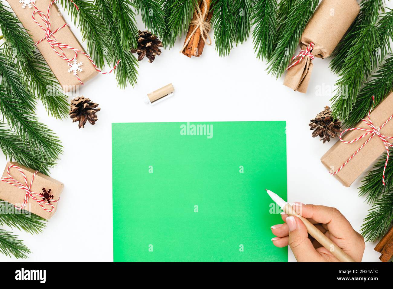Biglietto di Natale, lettera verde con spazio copia e una mano donna con penna artigianale su tavola bianca e ornamenti fatti di rami di pino con regali Foto Stock
