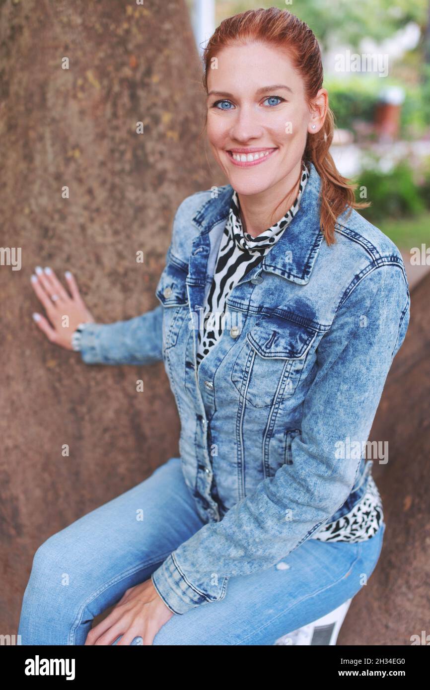 Giovane donna caucasica positiva di testa rossa che posa ad albero in parco in autunno Foto Stock