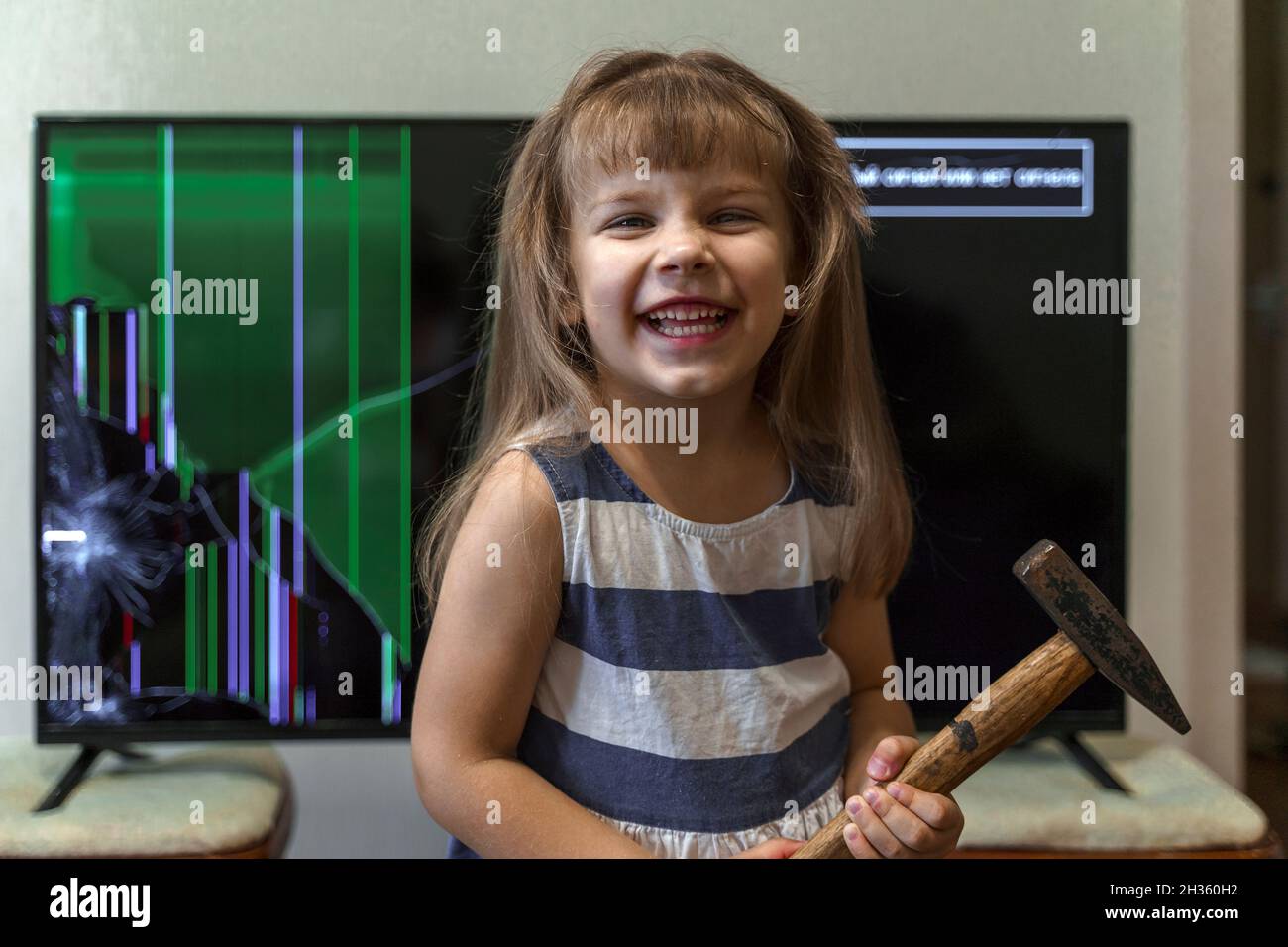 Carina bambina in piedi davanti alla tv con schermo rotto con martello Foto Stock