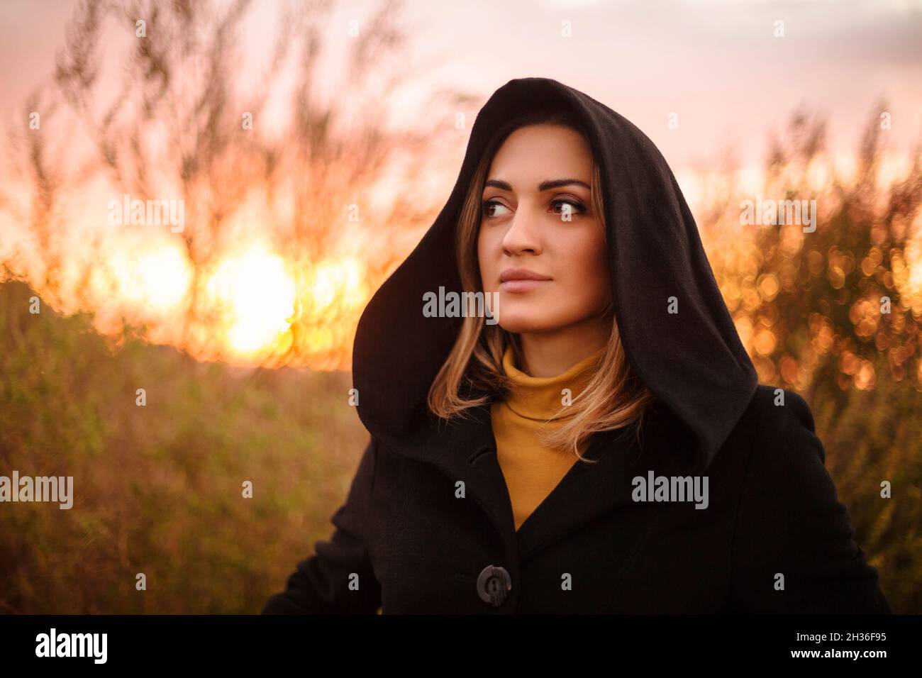 Giovane donna misteriosa in cappa nera guardando da parte mentre in piedi in autunno natura al tramonto, mystifying femmina camminare in prato in sera luce del sole Foto Stock