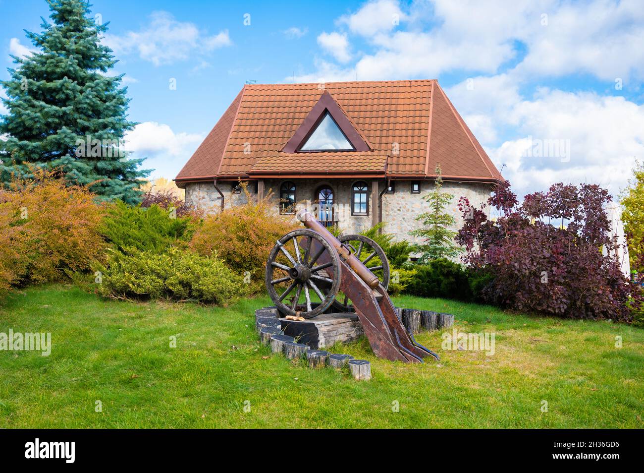 Vecchia casa in pietra con tetto rosso. Casa di campagna, casa padronale con prato e alberi, cannone vecchio come decorazione nel cortile Foto Stock