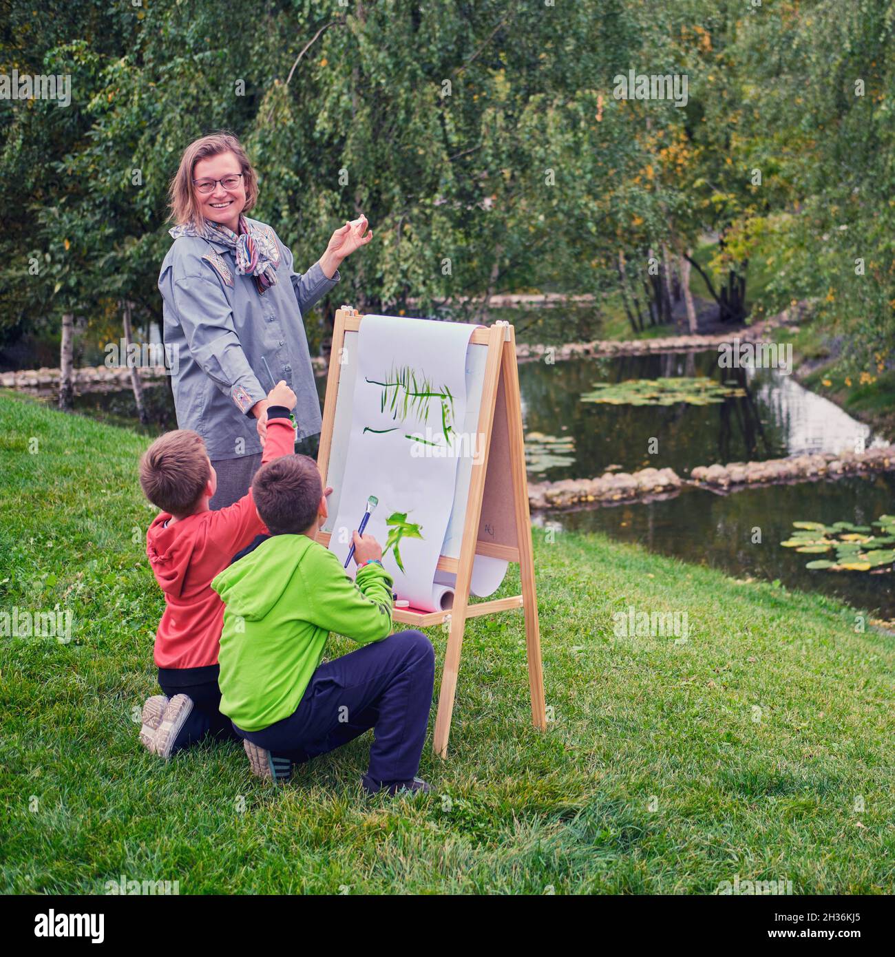 La madre insegna a dipingere con due allievi ragazzi. Donna insegnante artista dipinge con i bambini sulla carta natura e gli alberi vicino al fiume Foto Stock