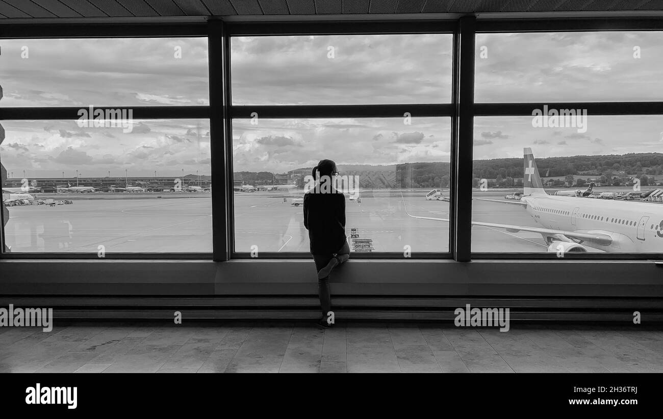 Scala di grigi di una giovane donna che guarda un aeroplano da una finestra di vetro Foto Stock