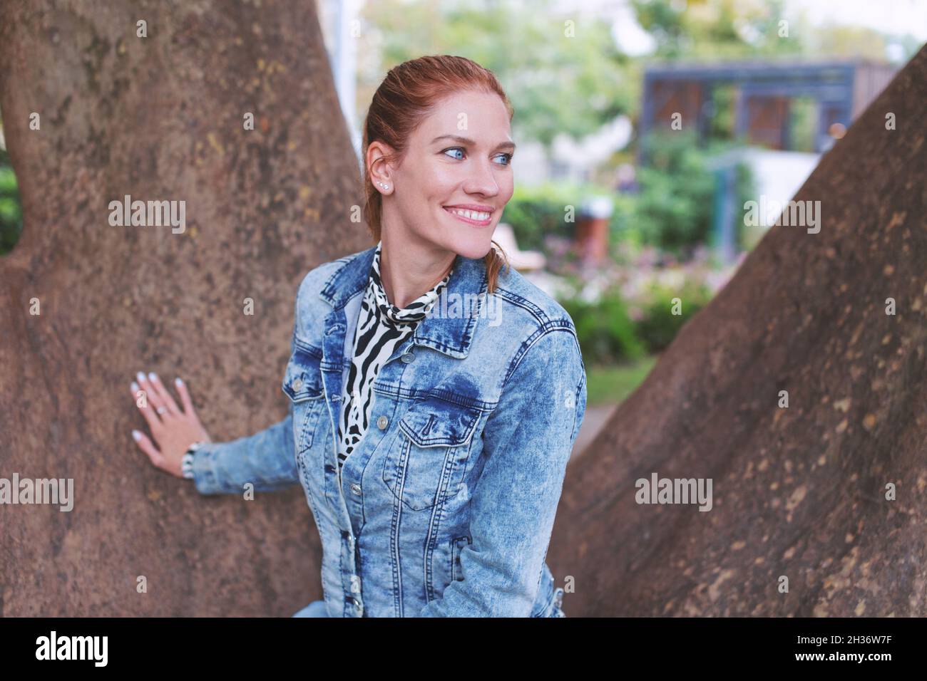 Giovane donna caucasica positiva di testa rossa che posa all'albero nel parco in autunno, guardando via Foto Stock