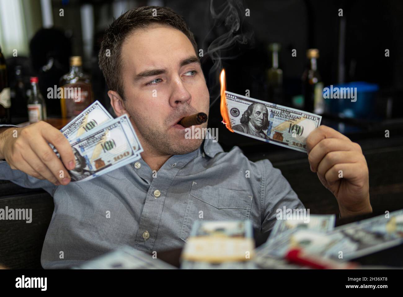 Uomo che illumina il suo sigaro con banconote da 100 dollari sullo sfondo delle bottiglie nel bar. Concetto di ricchezza e stravaganza. Foto Stock