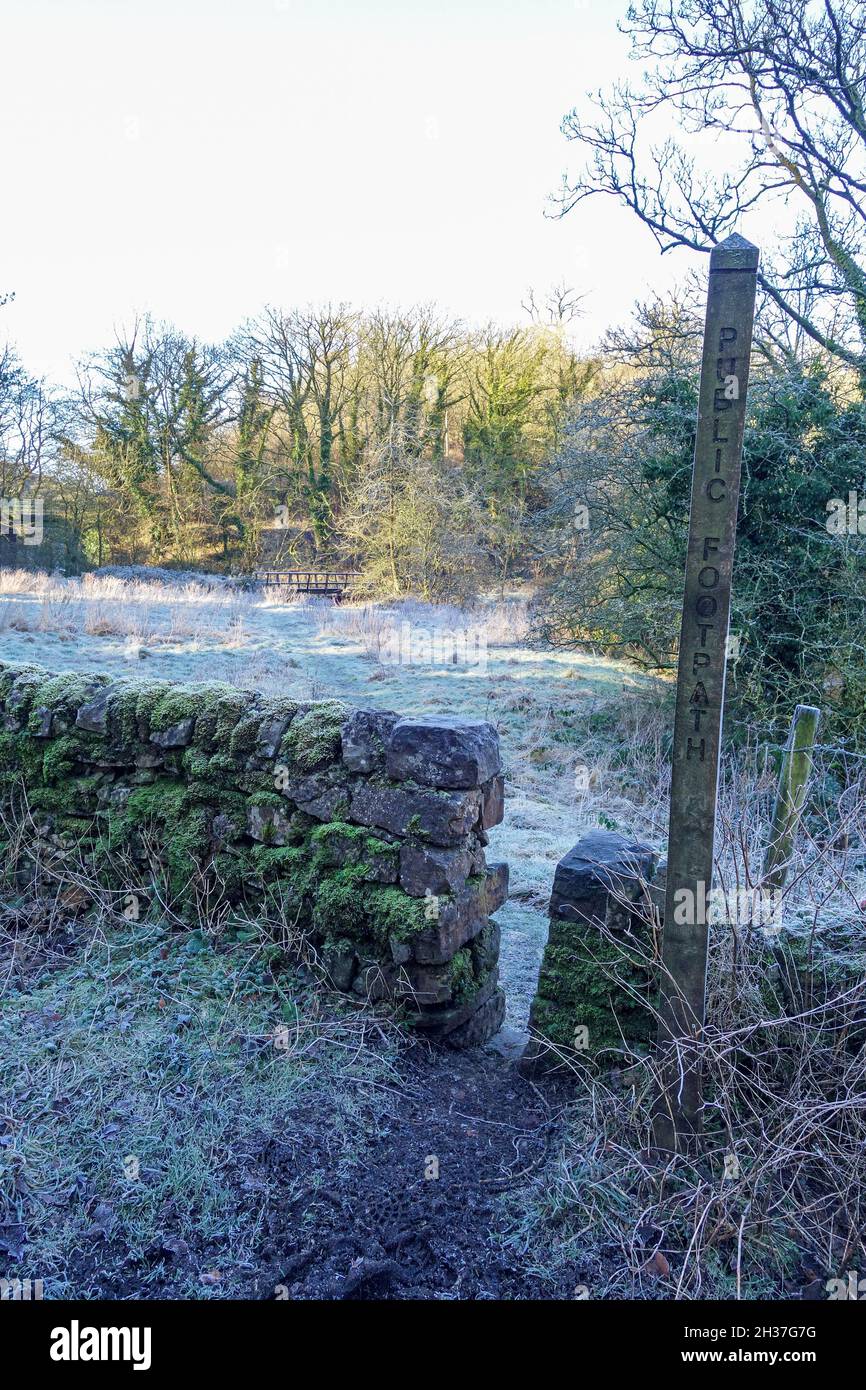 Un segnavia di legno e stiglia di pietra con il percorso che conduce ad un ponte di legno attraverso un campo coperto di gelo, Inverno, Staffordshire, Inghilterra, Regno Unito Foto Stock