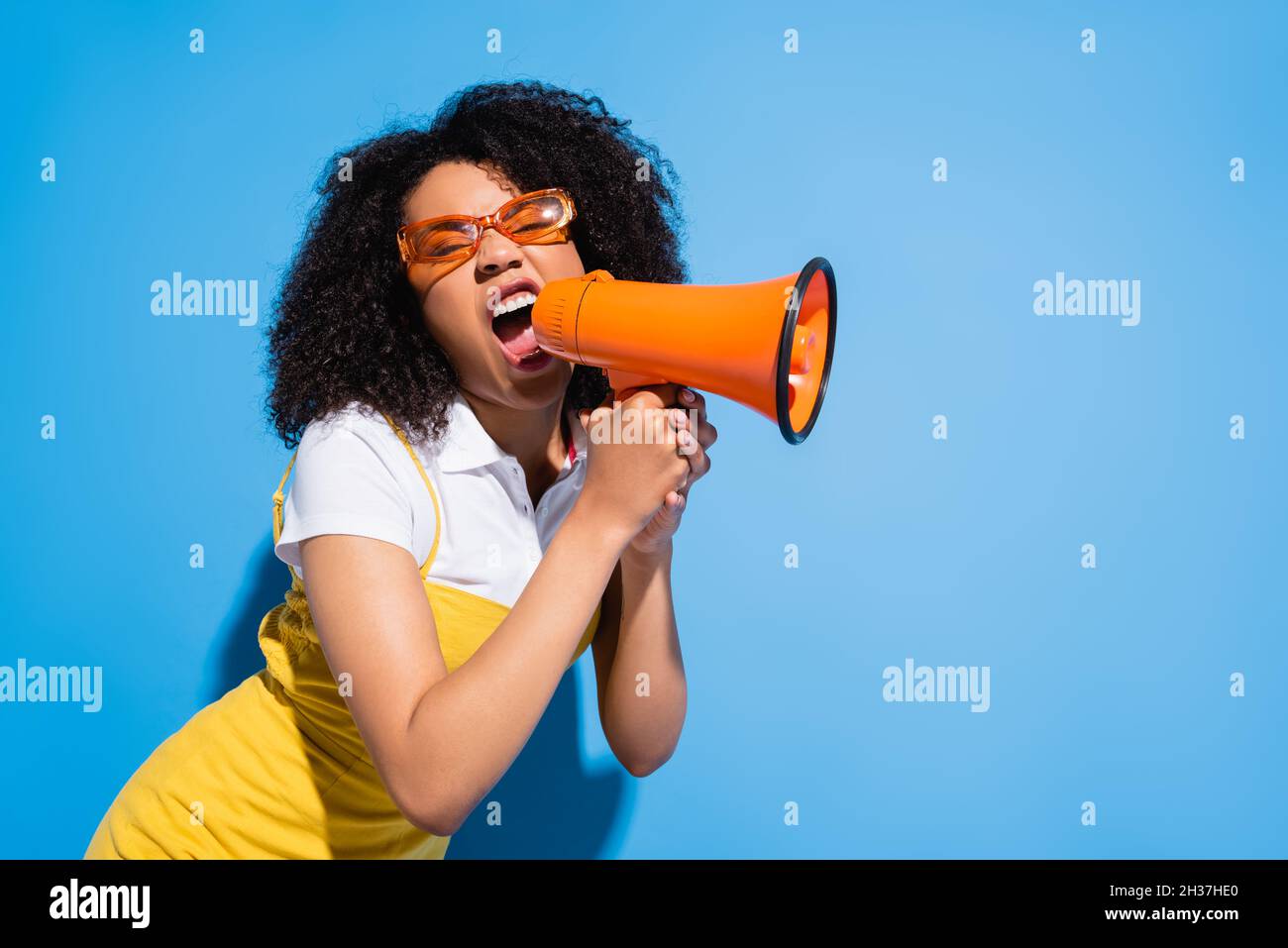 agitato afroamericana donna in occhiali trendy urlando in megaphone su blu Foto Stock